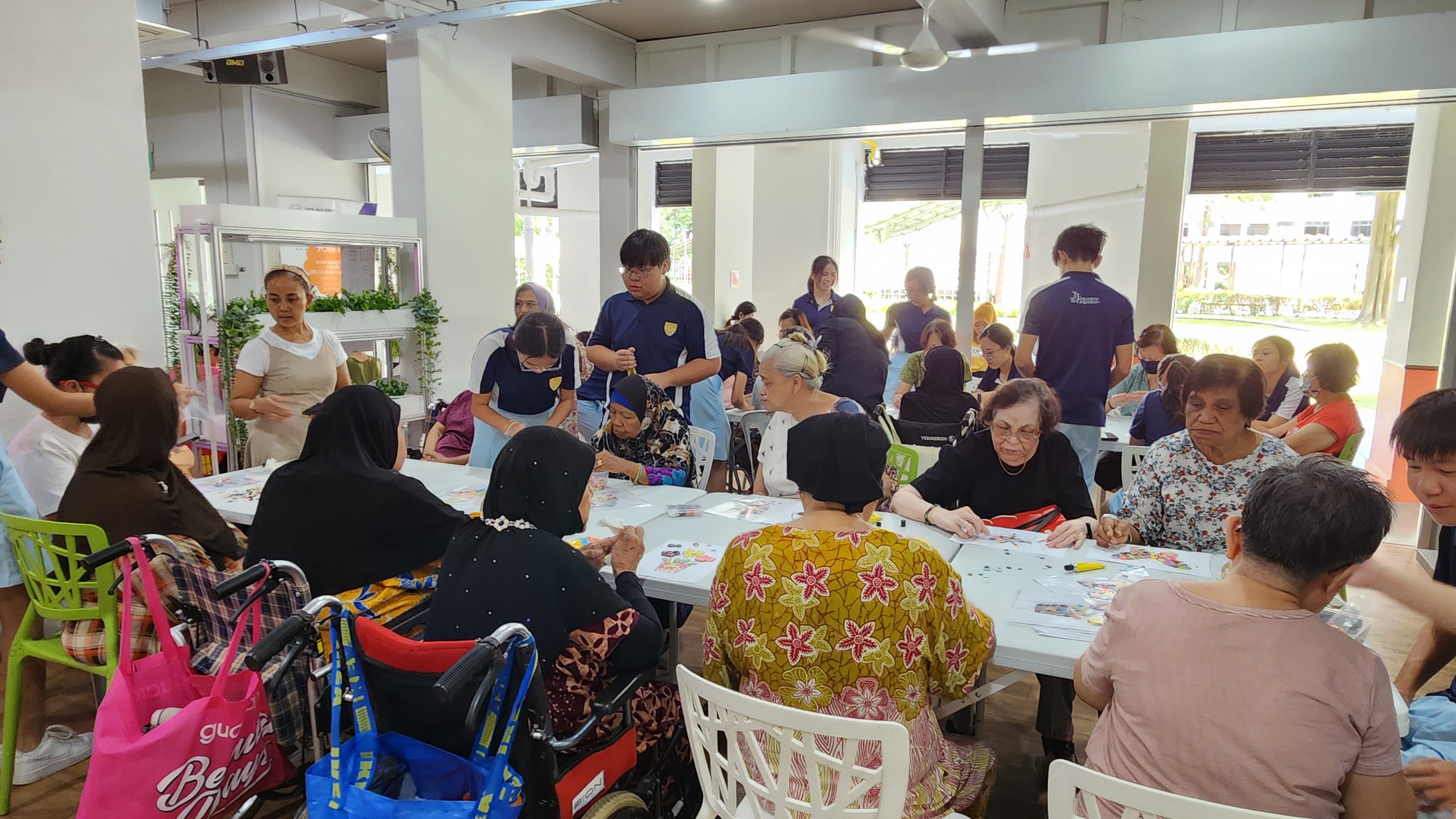 A large group of elderly people and young volunteers doing a craft activity together at tables in a bright indoor hall.